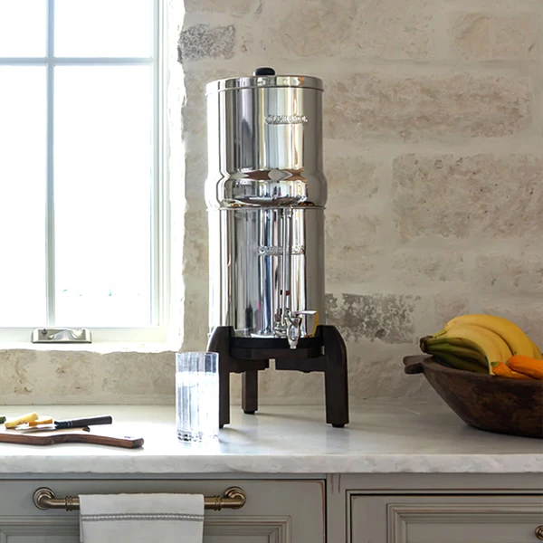 Woder Clearbrook 3.25 Gallon large countertop gravity water filter on wooden stand in modern kitchen with glass of water and fruit bowl