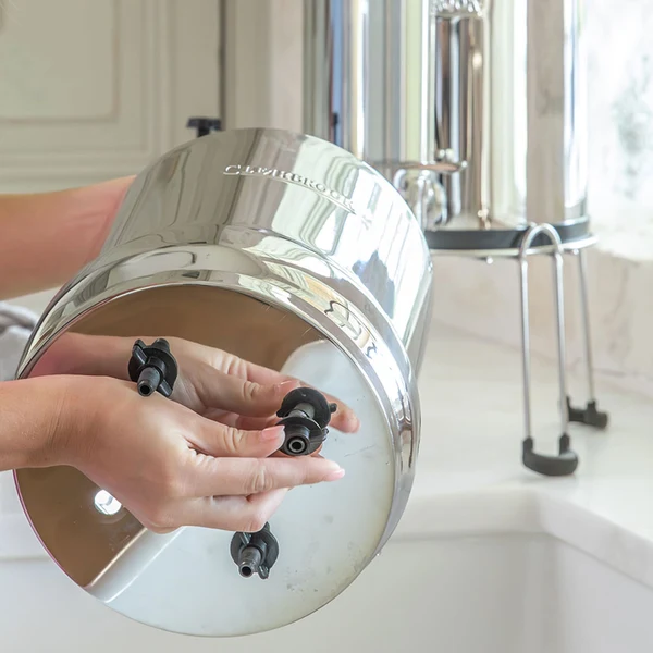 Hands installing CB9 filter elements into the upper chamber of Woder Clearbrook high flow countertop gravity water filter showing four filter ports on bottom of stainless steel vessel